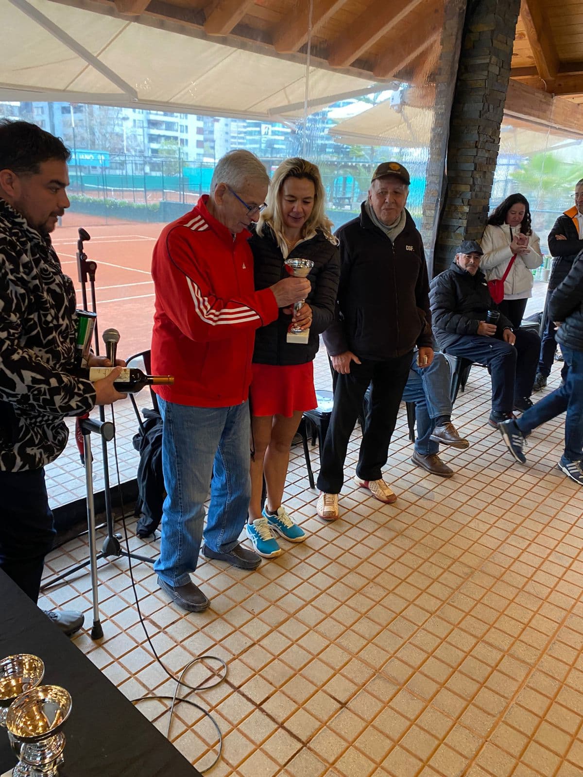 Man in red jacket and woman holding silver trophy at tennis award ceremony.