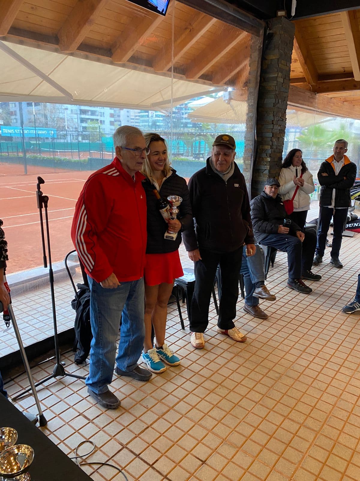 Woman holding a trophy and wine bottle poses with two men overlooking clay tennis courts.