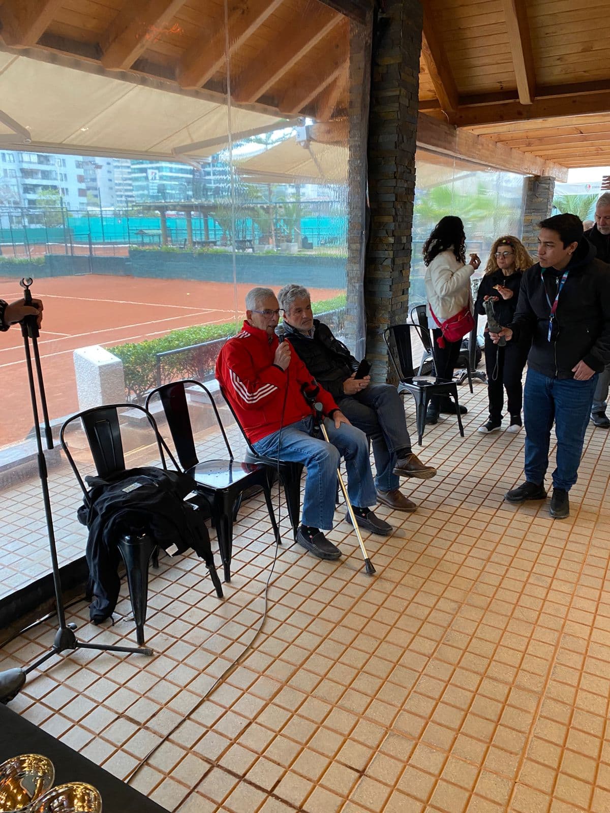 Older man in a red jacket speaks into a microphone overlooking red clay tennis courts.