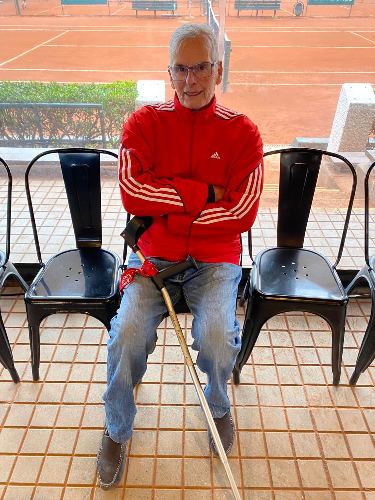 Elderly man in red tracksuit sits with a crutch, overlooking a clay tennis court.